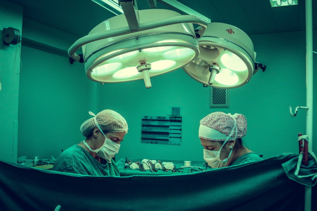 Zwei Chirurgen stehen in einem OP-Saal und operieren. Man sieht nur ihren Körper bis zur Schulter, davor hängt ein Tuch.