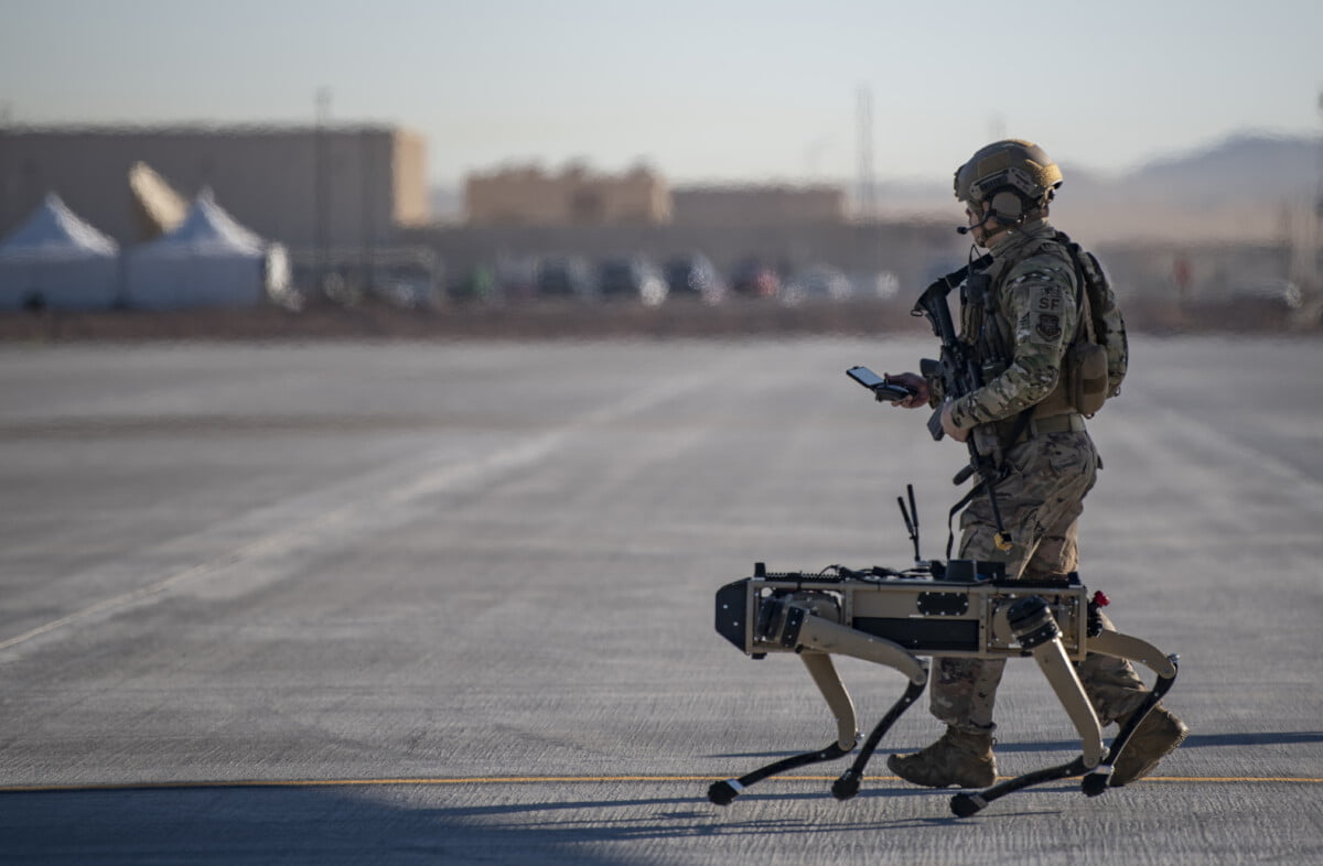 Ein Soldat läuft auf einem Asphaltweg, neben ihm läuft ein Roboter, der ähnlich aussieht wie ein Hund. Er hat vier Beine. Der Soldat steuert den Roboter über eine Ferbendienung, die er vor sich in der Hand hält.