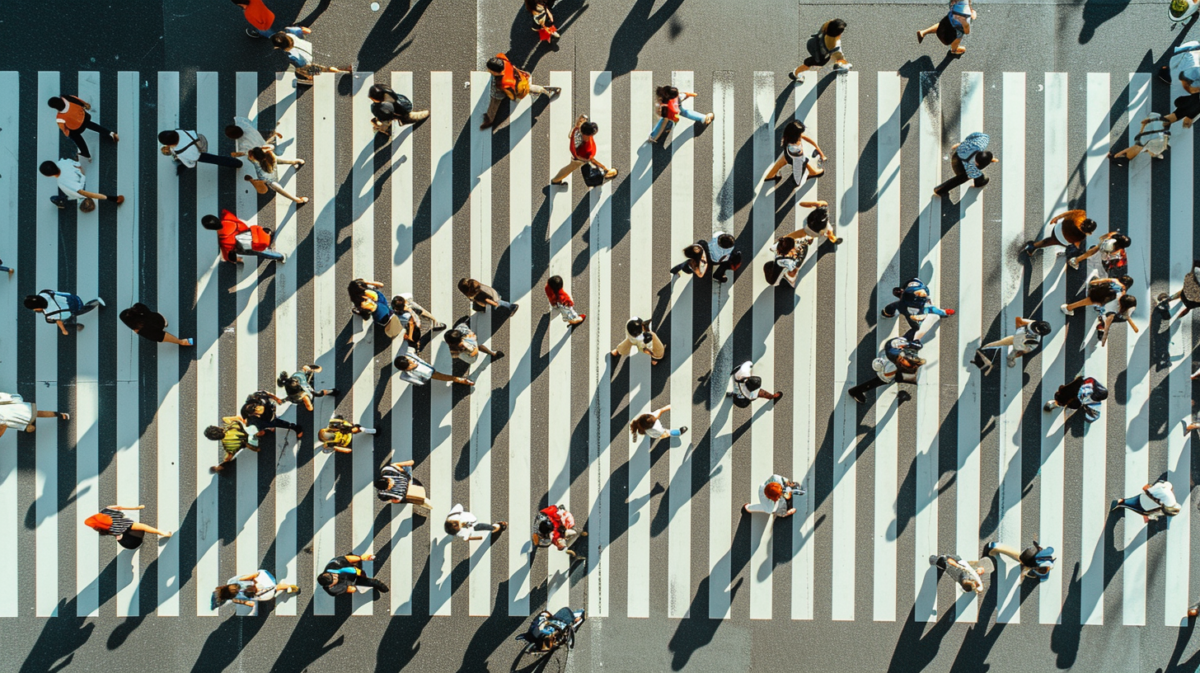 The image depicts a busy pedestrian crossing, where numerous people are walking in different directions. The photo appears to be taken from an elevated or aerial perspective, giving a top-down view of the scene. The crossing is marked by white stripes on the road, and the individuals are casting shadows, suggesting the time of day could be when the sun is at an angle, either morning or afternoon. The people are dressed in various colors, but muted tones predominate, with occasional bright colors like red and orange standing out. The image has a somewhat blurred or painted quality to it, possibly due to digital manipulation, creating an effect that is more artistic than a straightforward photograph