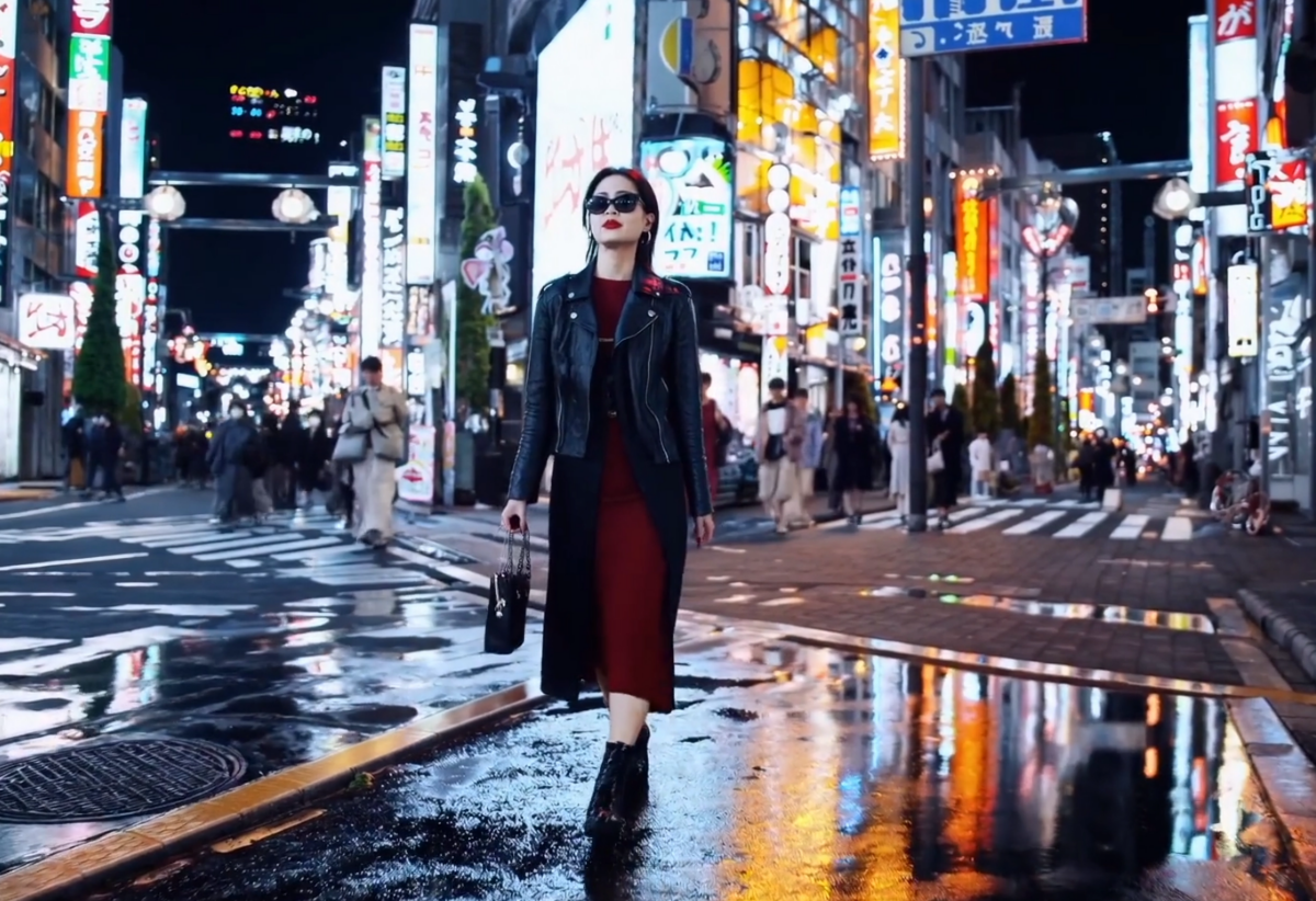 A stylish woman walks down a Tokyo street filled with warm glowing neon and animated city signage. She wears a black leather jacket, a long red dress, and black boots, and carries a black purse. She wears sunglasses and red lipstick. She walks confidently and casually. The street is damp and reflective, creating a mirror effect of the colorful lights. Many pedestrians walk about